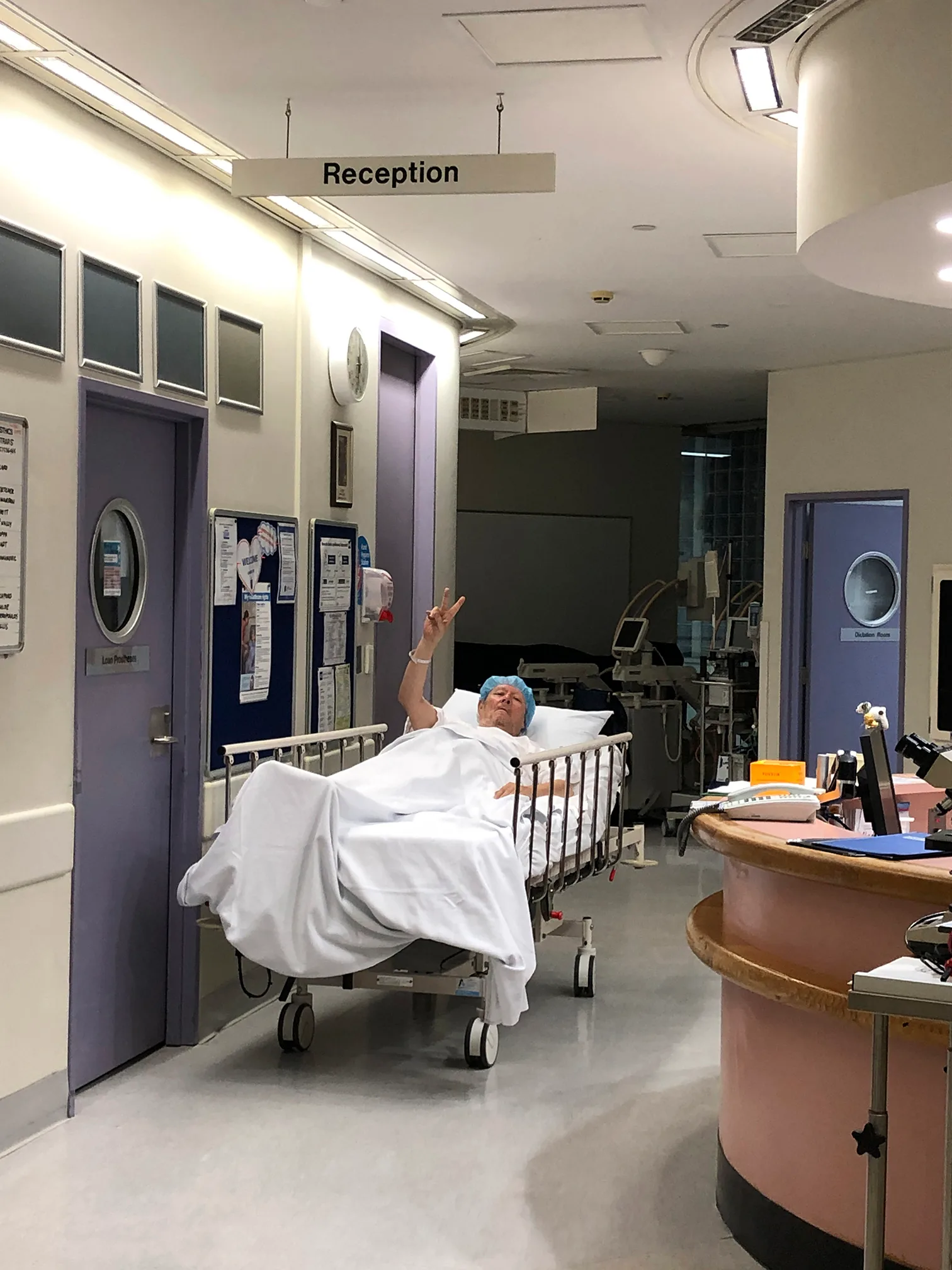 A man, wearing a blue surgical cap and covered in a white sheet, lies on guerney. He makes a peace sign as he is wheeled to surgery.