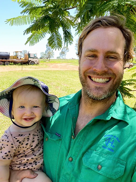 A man with sandy hair and beard, wearing a green shirt, holds a baby. The baby wears a floppy floral hat and a pale pink and black spotty t-shirt. Behind them are paddocks, trees and farm equipment.