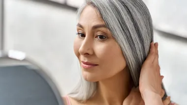 Portrait of a beautiful, happy middle-aged woman. She is touching her long grey hair, looking at herself in the mirror.