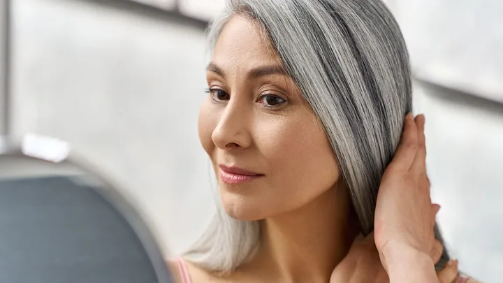 Portrait of a beautiful, happy middle-aged woman. She is touching her long grey hair, looking at herself in the mirror.