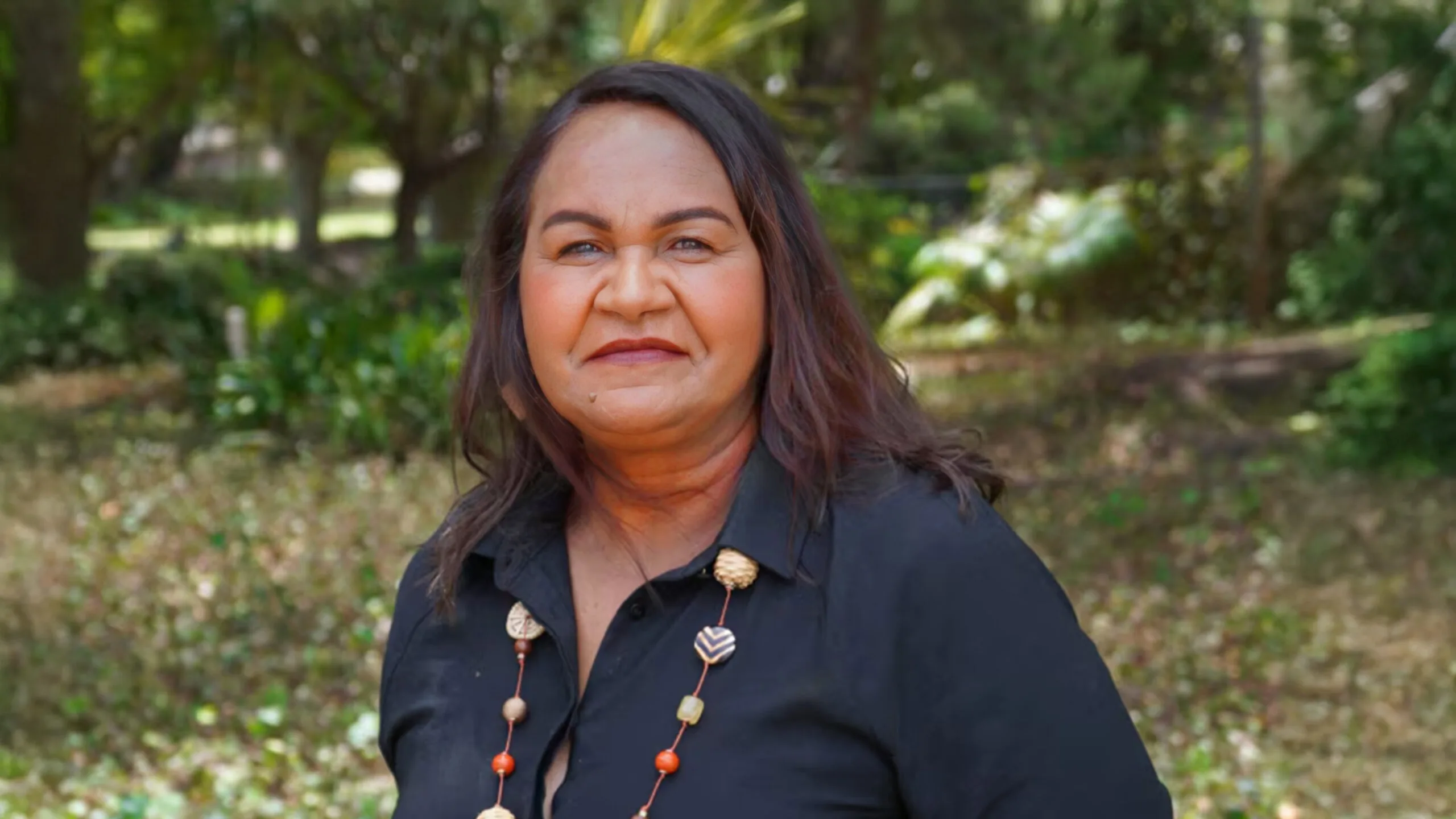 Donna Nelson, a woman in a navy shirt with a colourful necklace, stands in a forested area. She has long dark brown hair and a slight smile.
