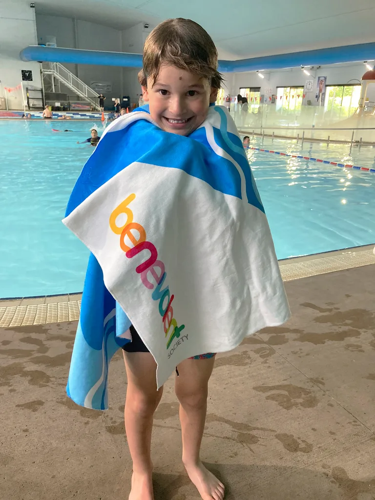 Young boy, George, stands by a swimming pool wrapped in a towel