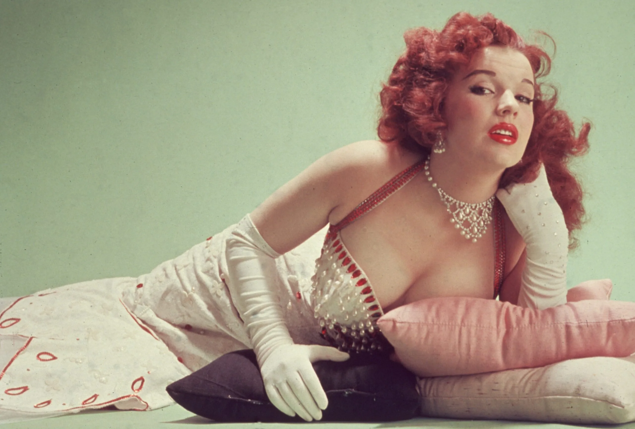 circa 1955: Studio portrait of Blaze Starr, reclining on pillows on the floor, wearing a red-trimmed white dress and white elbow-length gloves. (Photo by Hulton Archive/Getty Images)