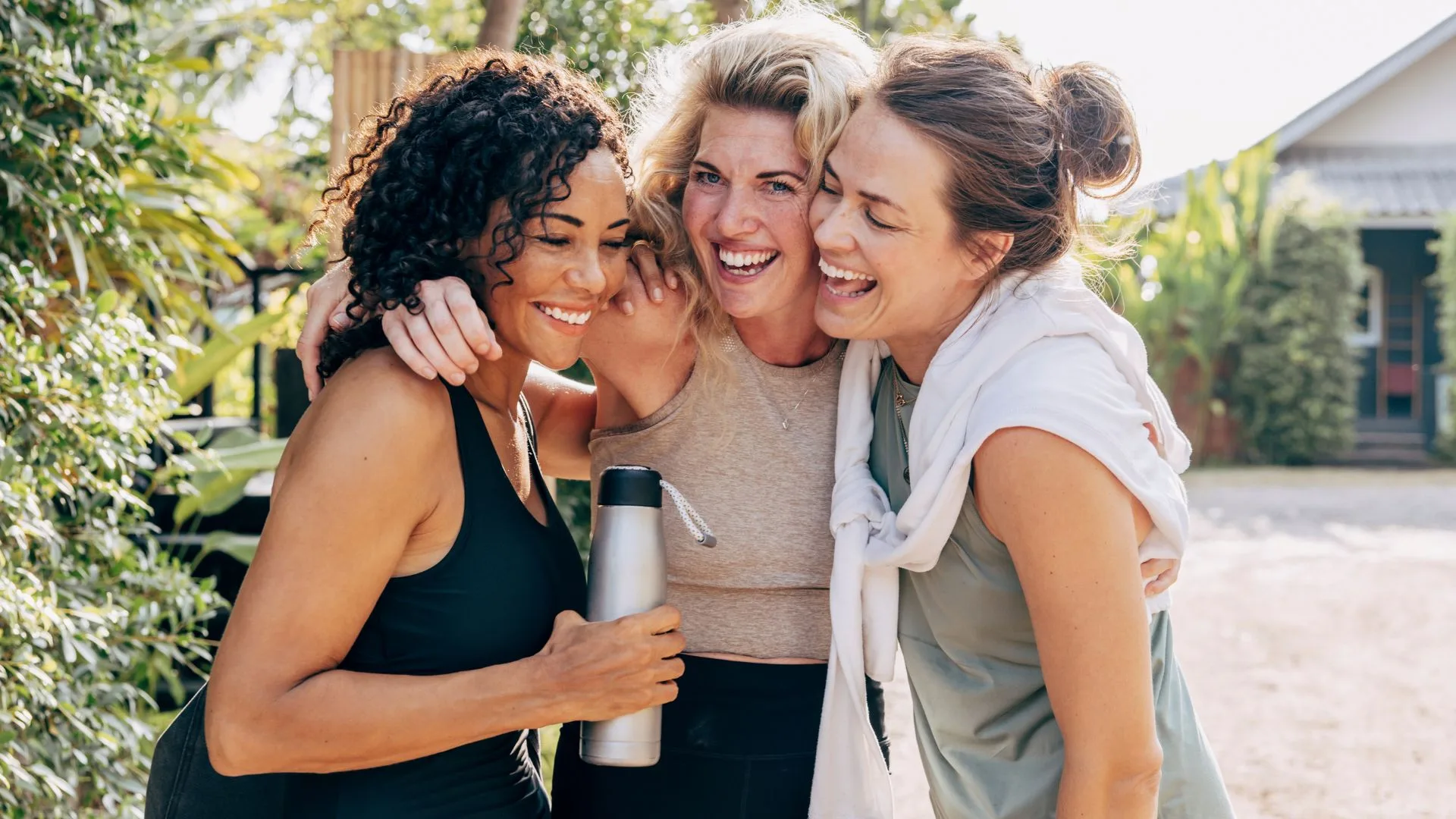 Three women smiling and embracing outdoors after a workout, appearing relaxed and happy in casual activewear.