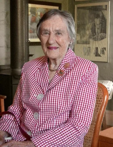 Jessie as an older woman wears a red and white checked blazer and her gold medal of recognition on the lapel. Celebrating our servicepeople on Anzac Day