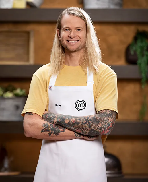 Blonde man with tattoos, wearing a yellow shirt and "Pete" MasterChef apron, smiling with arms crossed.