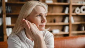 A close-up of a middle-aged woman with blonde hair. She is resting her chin on her hands, looking stressed, sad, tired and exhausted. She is sitting on a brown leather couch in front of a bookshelf in an office.