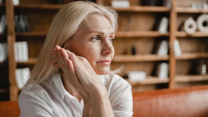A close-up of a middle-aged woman with blonde hair. She is resting her chin on her hands, looking stressed, sad, tired and exhausted. She is sitting on a brown leather couch in front of a bookshelf in an office.