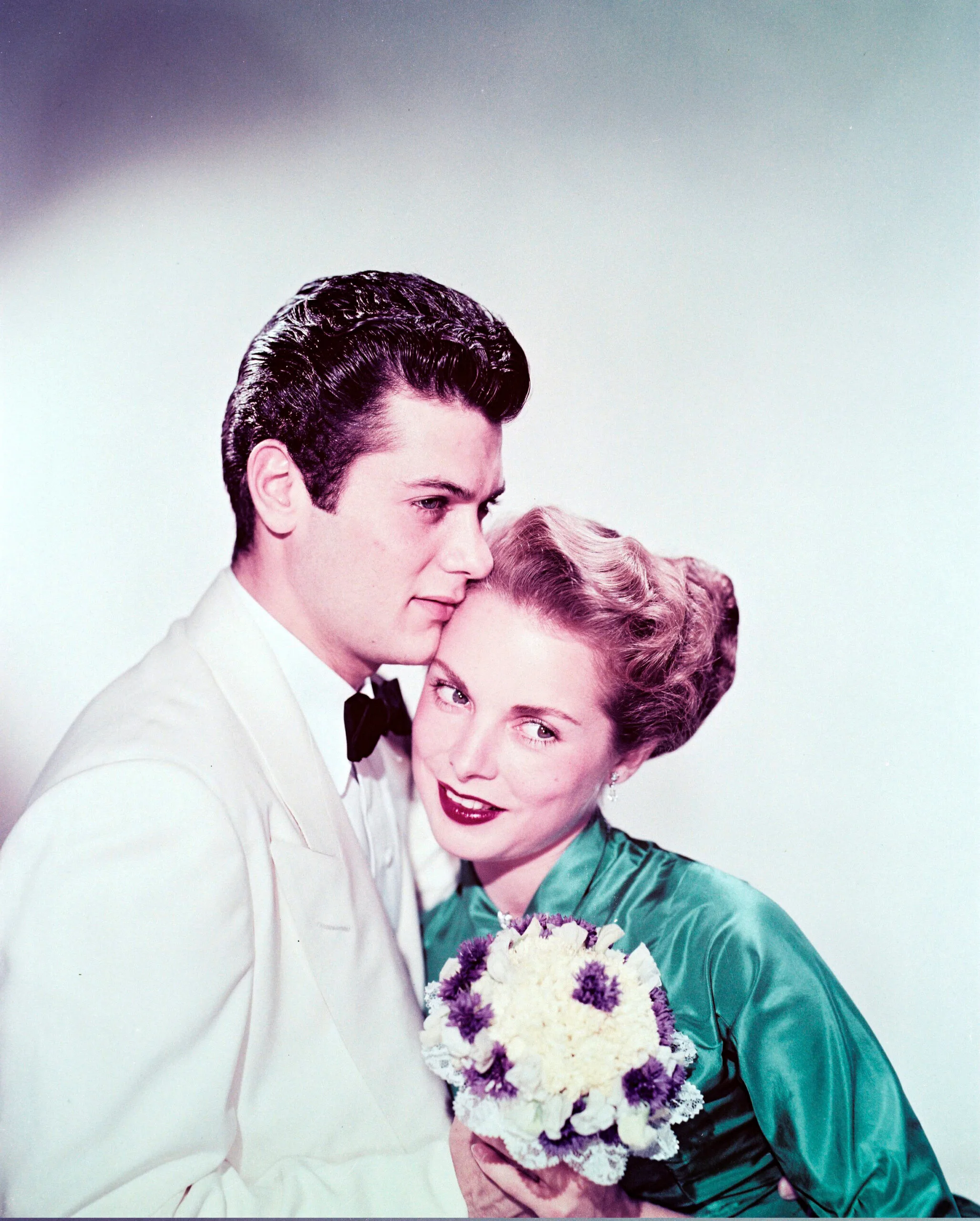 Tony Curtis (1925-2010), US actor, wearing a white dinner jacket with a white shirt and a black bow tie, posing with his wife, Janet Leigh (1927-2004), US actress, who wears a turquoise blouse and holds a small bouquet of flowers in a studio portrait, against a white background, circa 1955. (Photo by Silver Screen Collection/Getty Images)