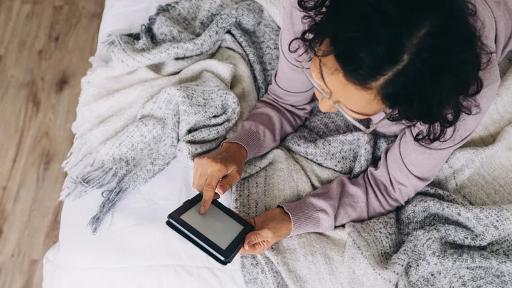 Woman with dark hair and glasses on bed reading an ereader