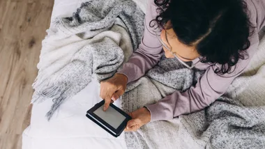 Woman with dark hair and glasses on bed reading an ereader
