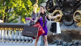 Blair Waldorf in the TV show, Gossip Girl, played by actress Leighton Meester. She is wearing a stylish purple dress holding multiple luxury shopping bags, including Gucci and Chanel, while standing on a decorative bridge.