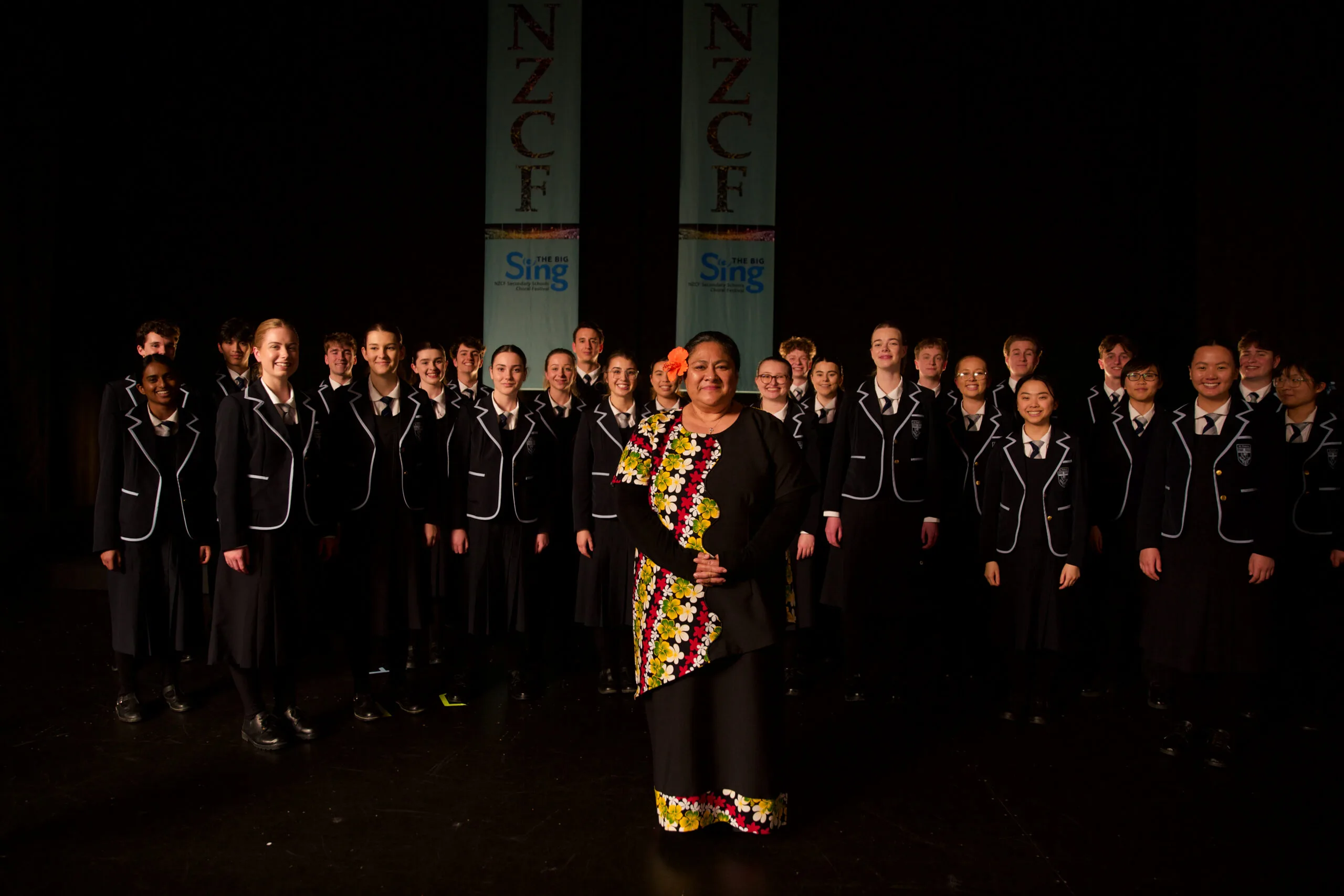 A full choir of teenage school children in formal uniform stand behind a woman wearing a very elegant, floor-length black Samoan dress with a diagonal white, red, yellow and green floral design.
