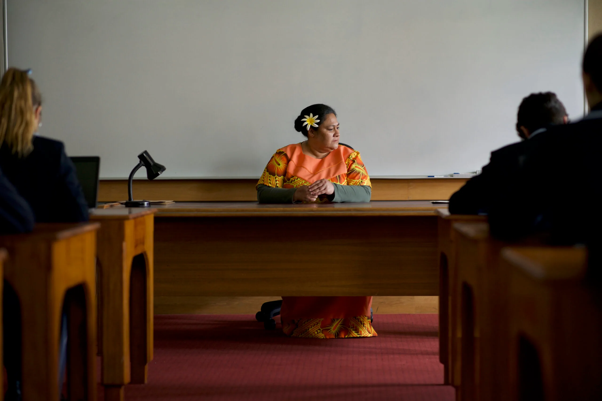 A woman in an orange Saman dress, wearing a flower in her hair, sits behind a teacher's desk.