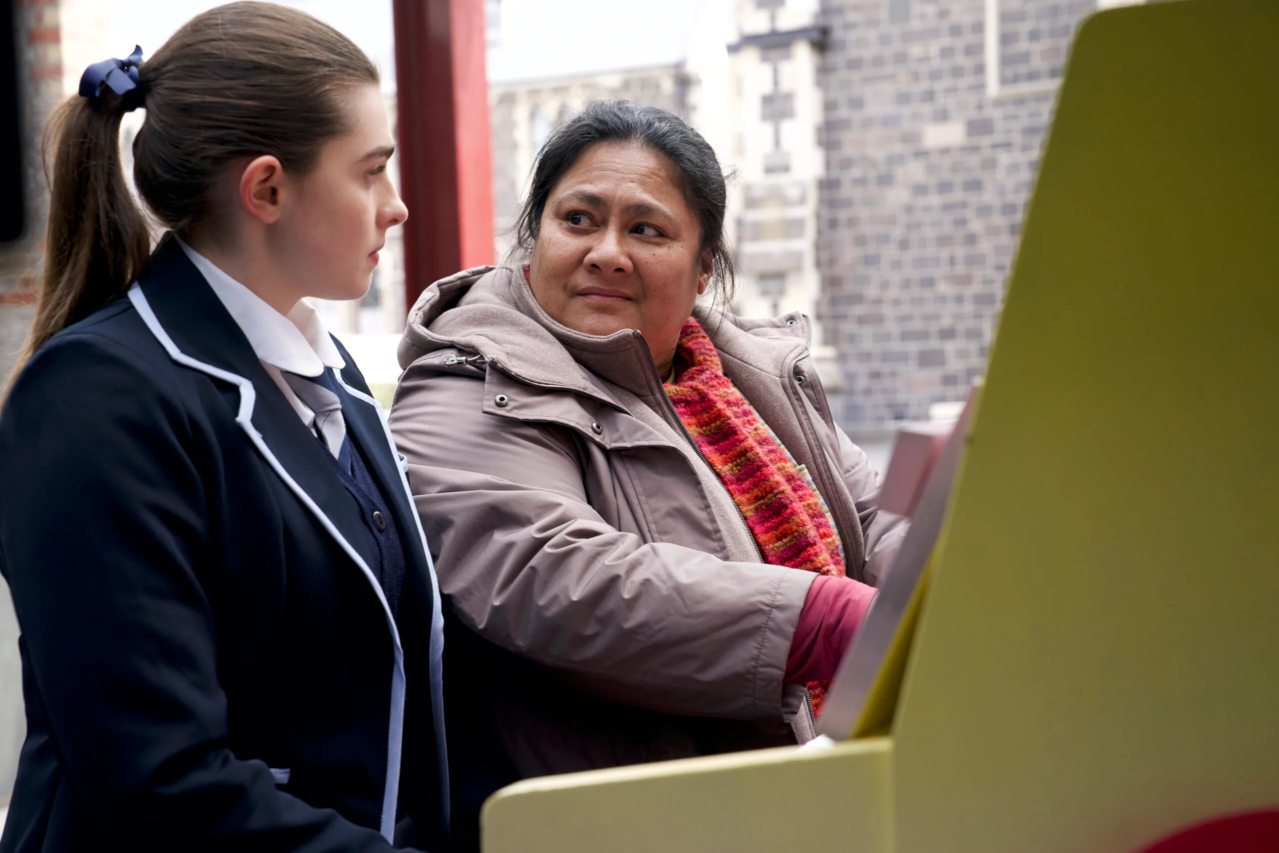 A schoolgirl in uniform sits beside an older woman at a piano in an outdoor playground.