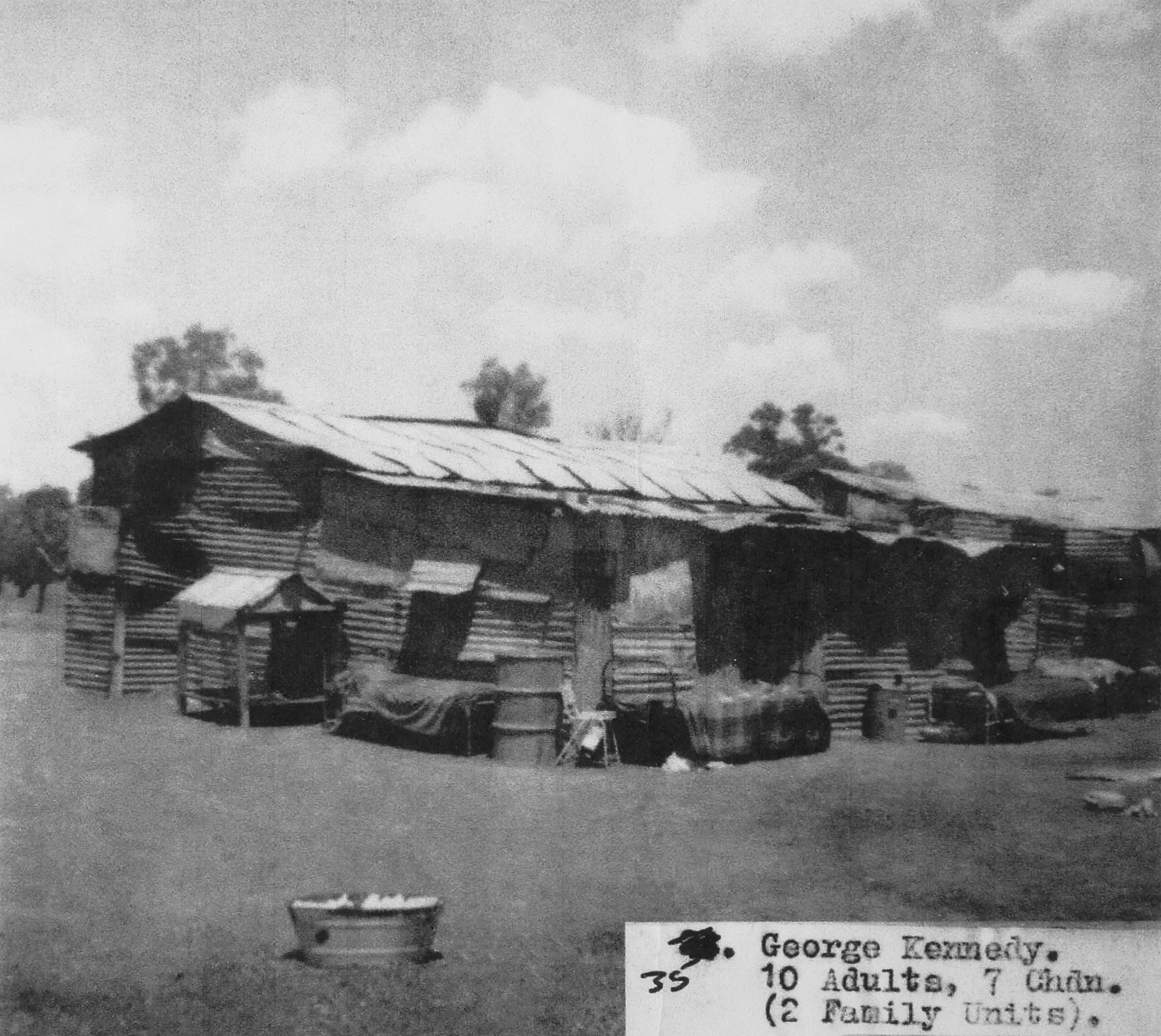 A black and white whotograph of a small corrugated iron home. Thre is a washing tub in the foreground.