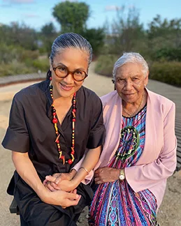 Kylie Kwoong wears round tortoiseshell glasses, a black pantsuit and a necklace in the Aboriginal flag's colours - red, black and yellow. She holds hands with Aunty Beryl who wears a colourful print dress and a pink jacket. They stand in a native food garden in Redfern.