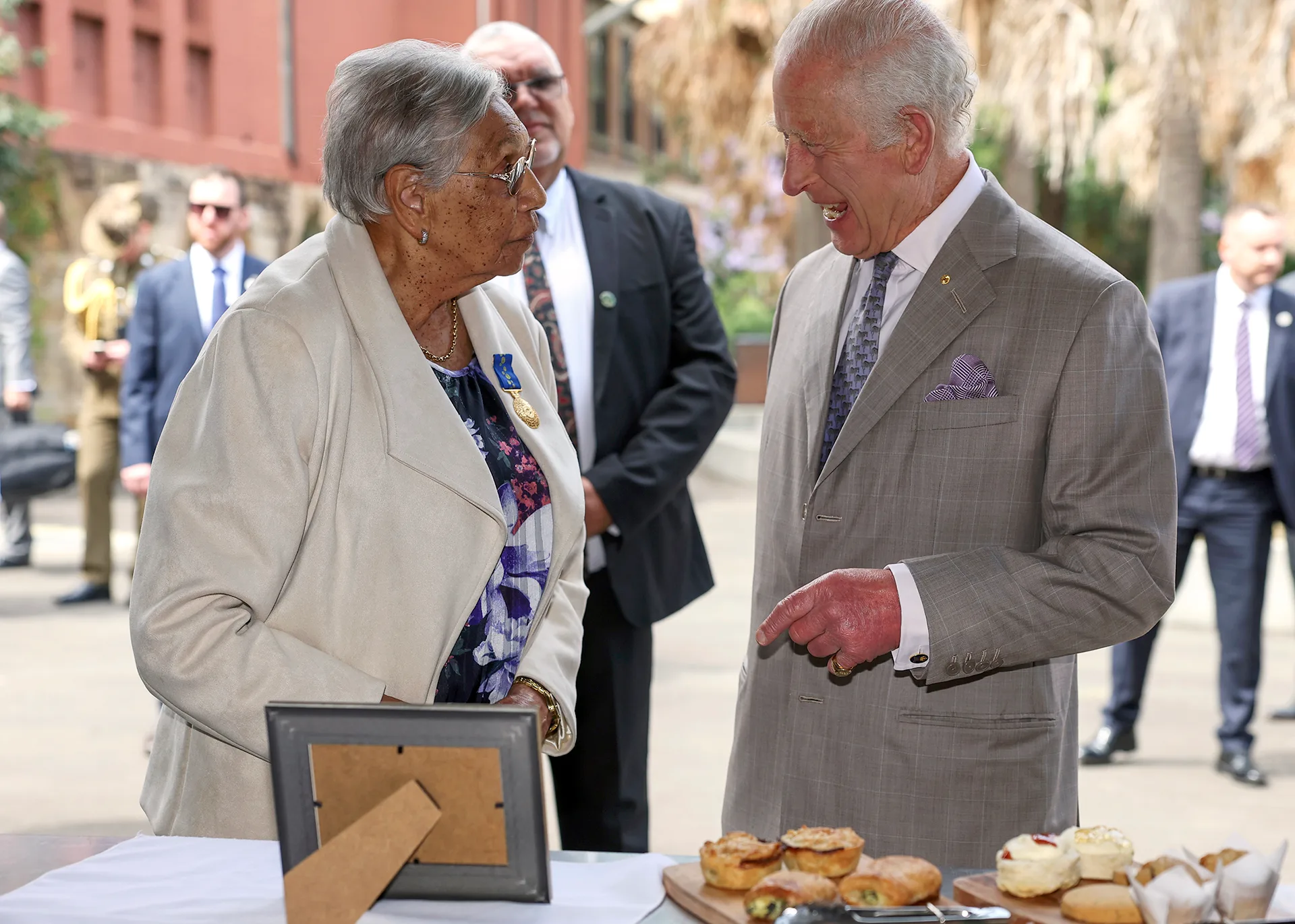 Aunty Beryl, wearing a white jacket and a floral dress, is in deep conversation with King Charles, dressed in a grey suit. In the foreground is a table of baked goods.
