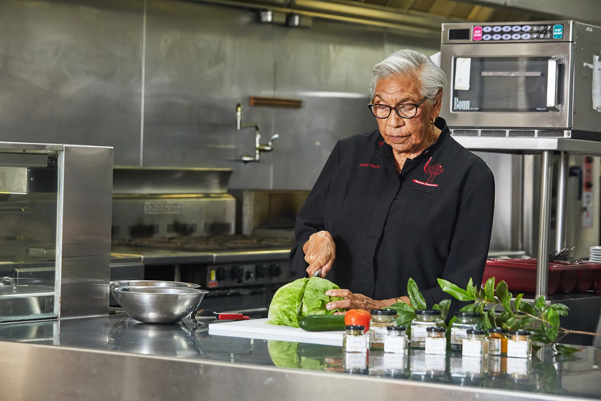 Aunty Beryl wears the navy uniform of her catering company and prepares a salad in her commercial kitchen.