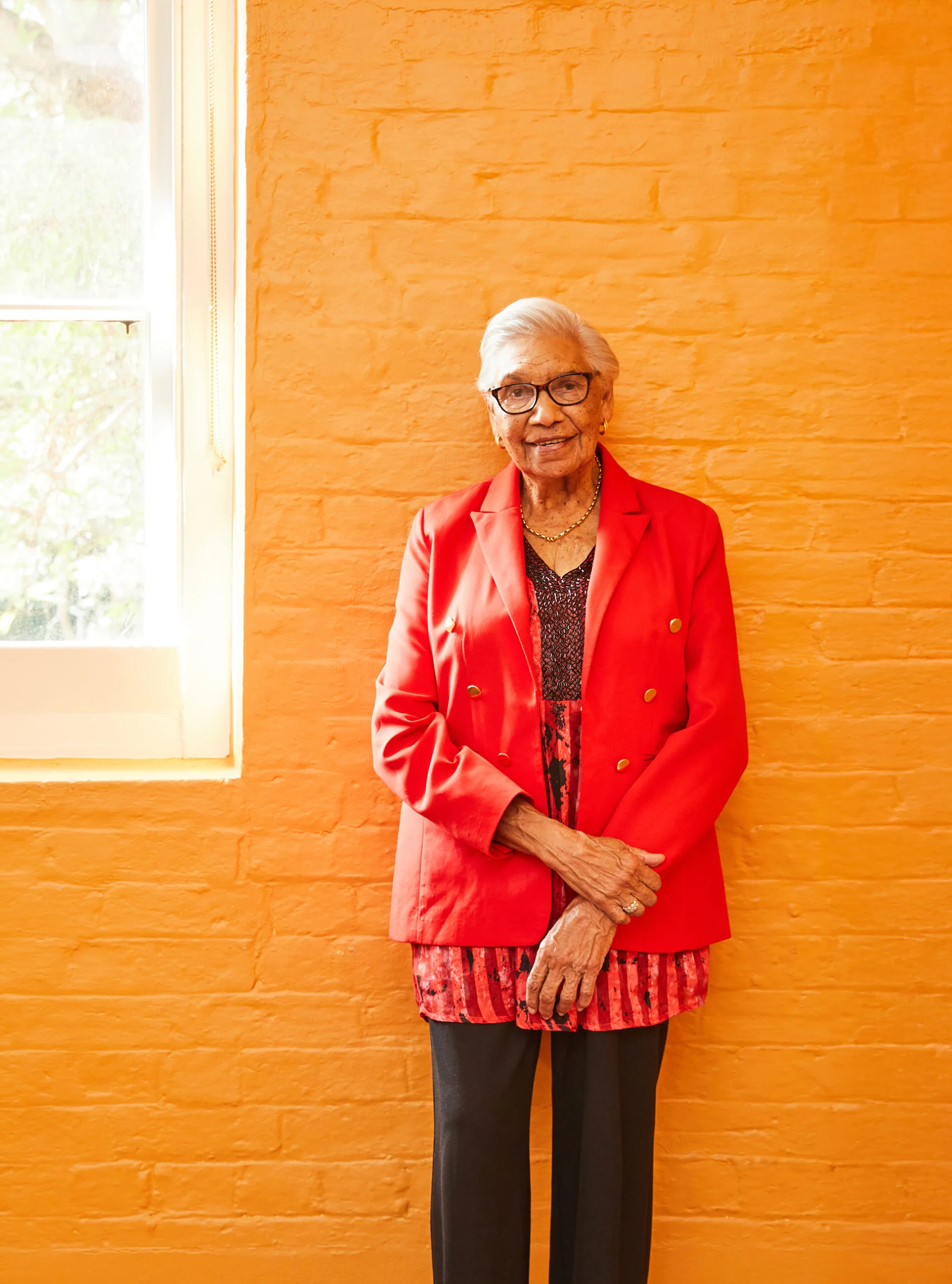 Aunty Beryl, standing against an orange wall, wears black trousers and a red jacket.