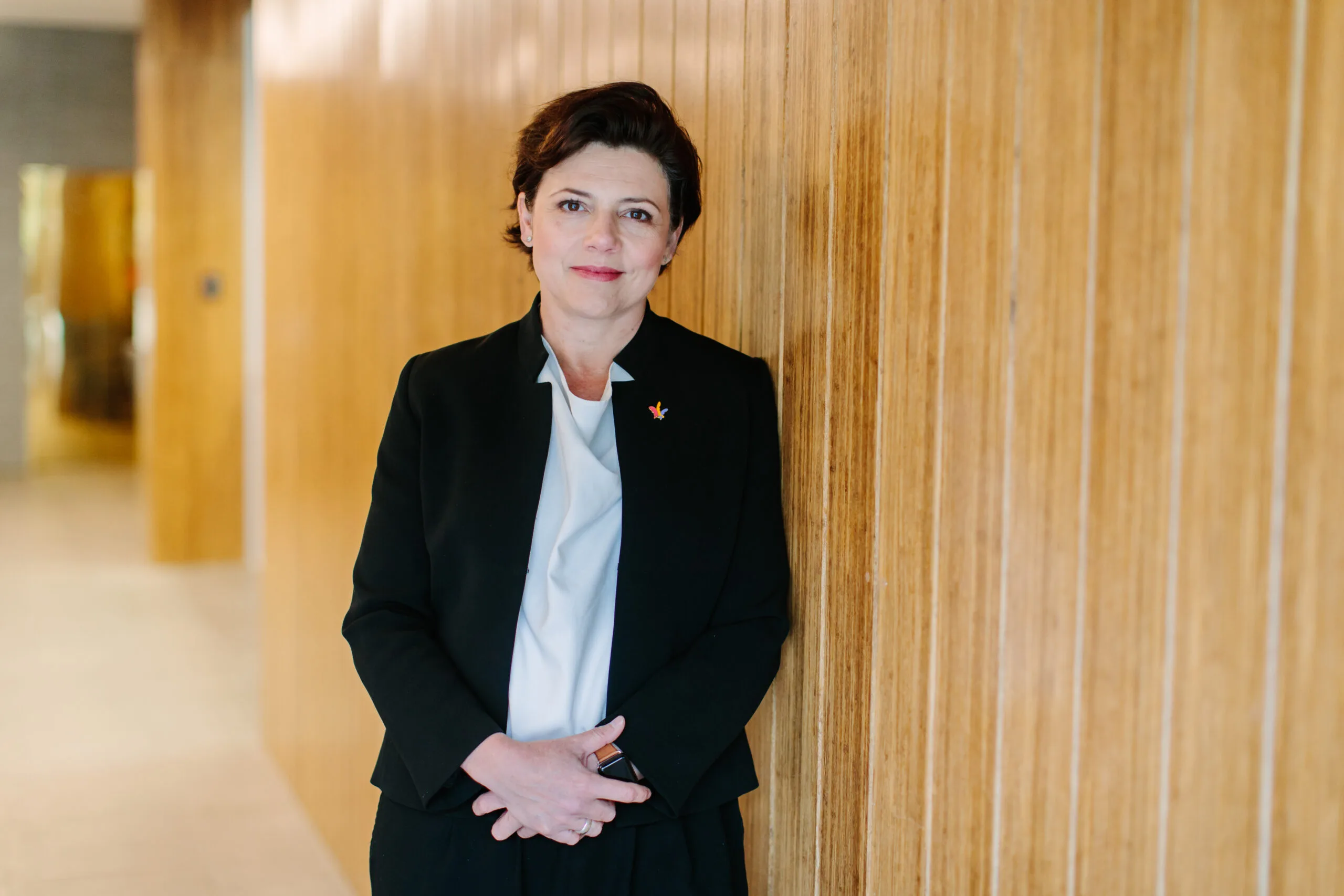 Georgie Harman wears a suit and a pale blue shirt. She stands in a wood-panelled corridor, smiling thoughtfully.