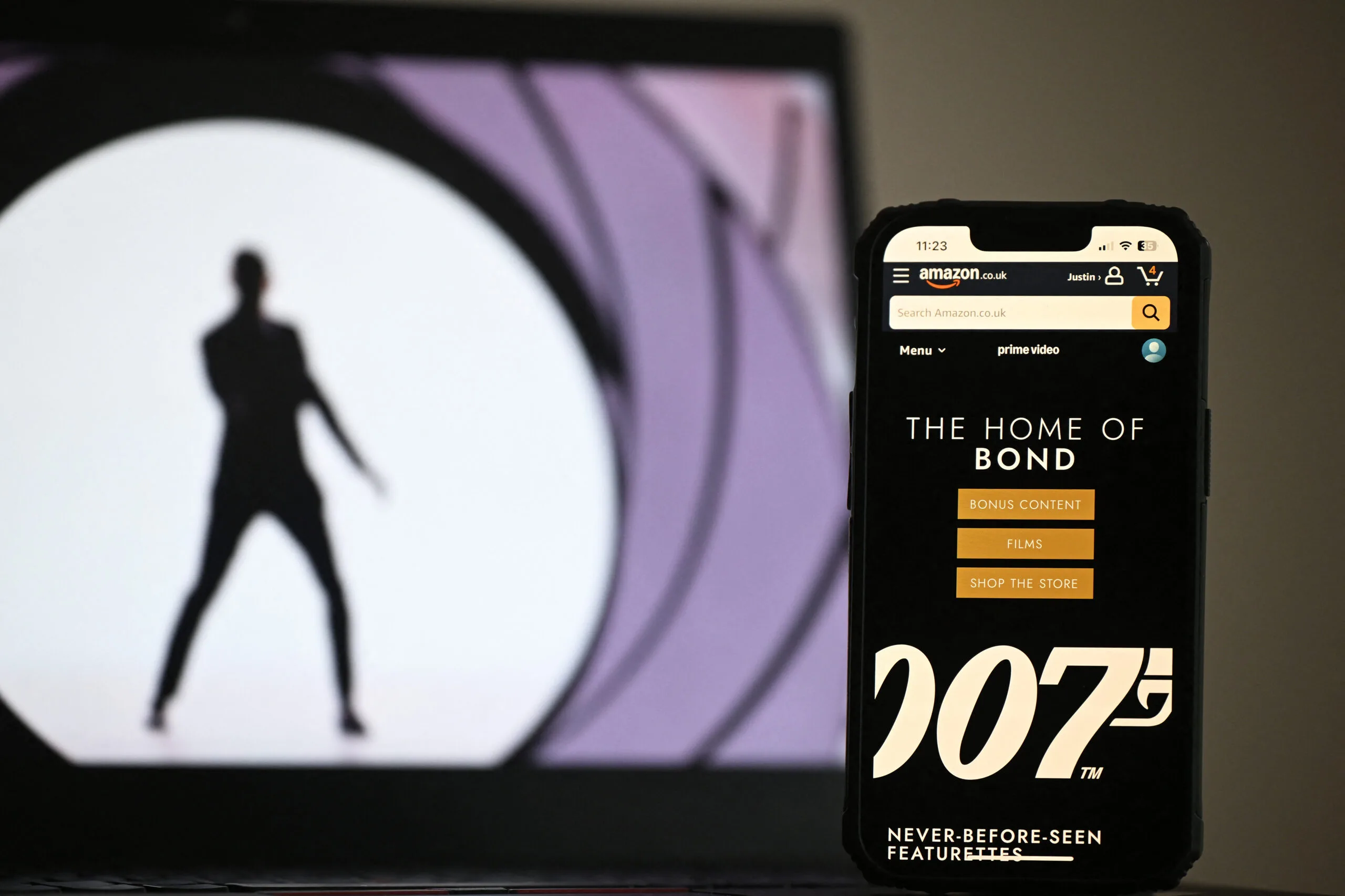 In this photo illustration created in London on February 23, 2025 the Amazon.co.uk app displays "The Home Of Bond" on a phone screen next to an photograph displayed on a laptop screen. 