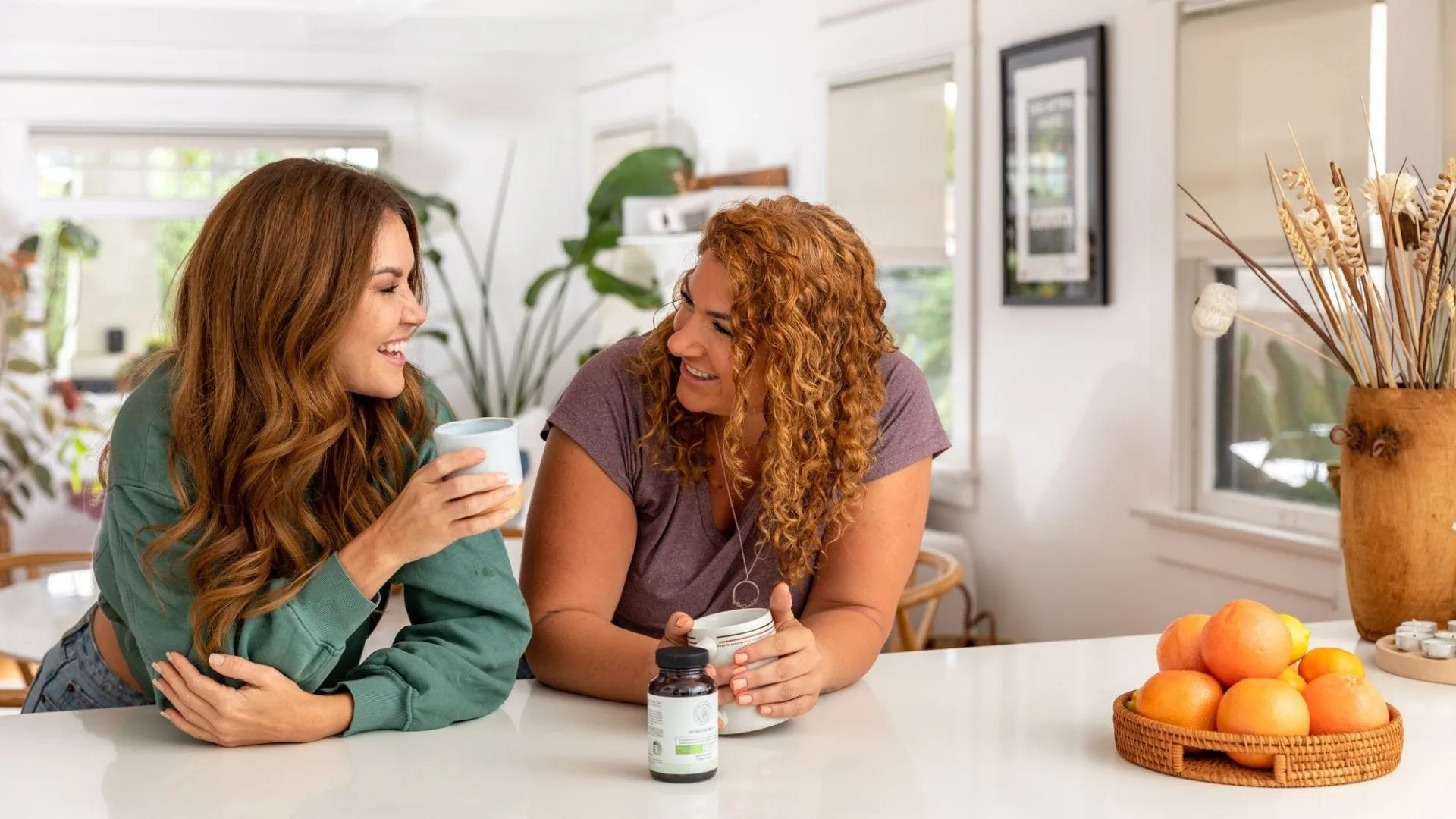 Two women smiling and talking in a bright kitchen, with one holding a mug and the other holding a jar of Happy Mammoth supplements.