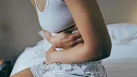 Woman sitting on a bed clutching her lower abdomen, appearing to experience bloating or stomach discomfort.