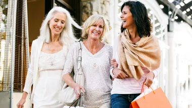 Three happy women, two blonde and one with dark hair, are smiling and walking together, carrying shopping bags. The woman on the right has an orange shopping bag.