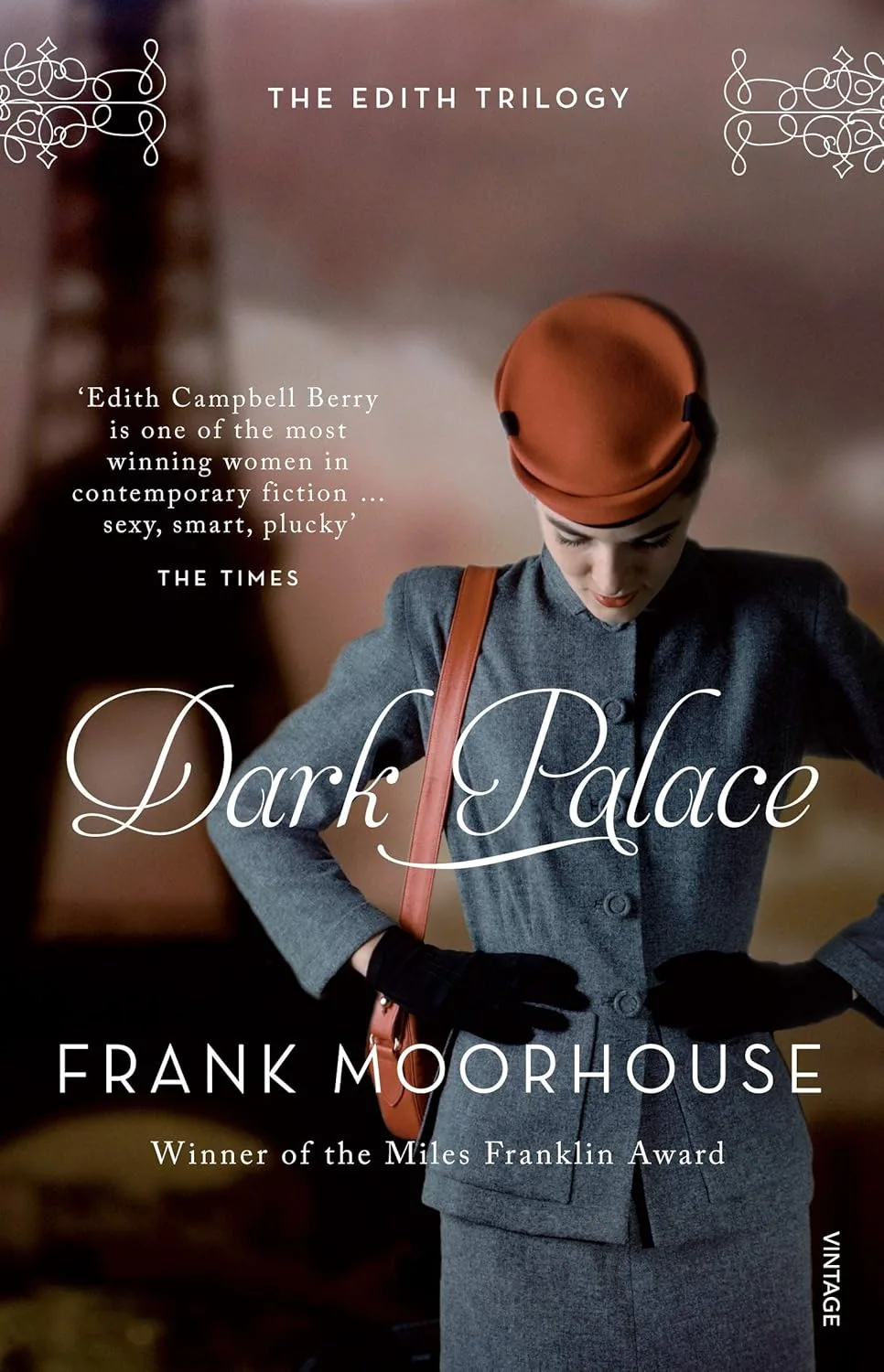 Book cover Dark Palace Frank Moorhouse. A woman in a dark grey jacket holds her hands on her hips, looking downward. She has a red hat and handbag.