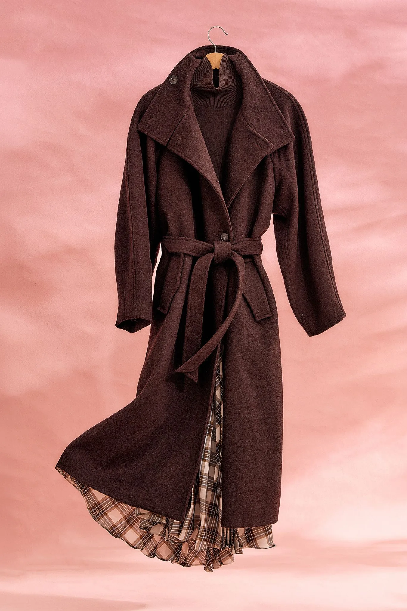 The Elka Collective Angelica Wool-Blend Coat in a rich chocolate brown, displayed on a wooden hanger against a textured pink studio backdrop. The long, belted trench coat is styled over a matching brown turtleneck knit and a flowing, pleated plaid skirt that peeks out from beneath the hem.