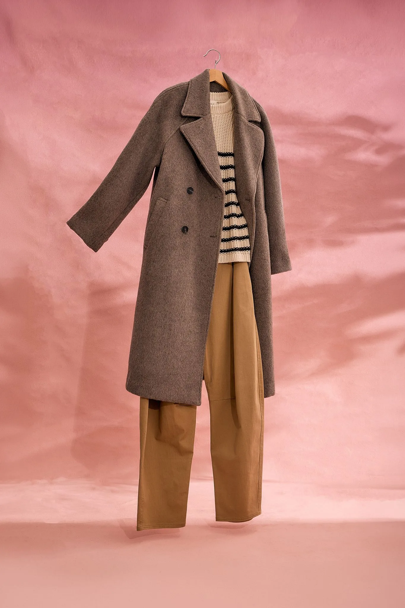 The Elka Collective Montana Wool-Blend Coat featured in a multi-panel layout on a soft pink background. The longline, structured coat is styled with a classic cream and black striped knit sweater and tailored tan trousers.