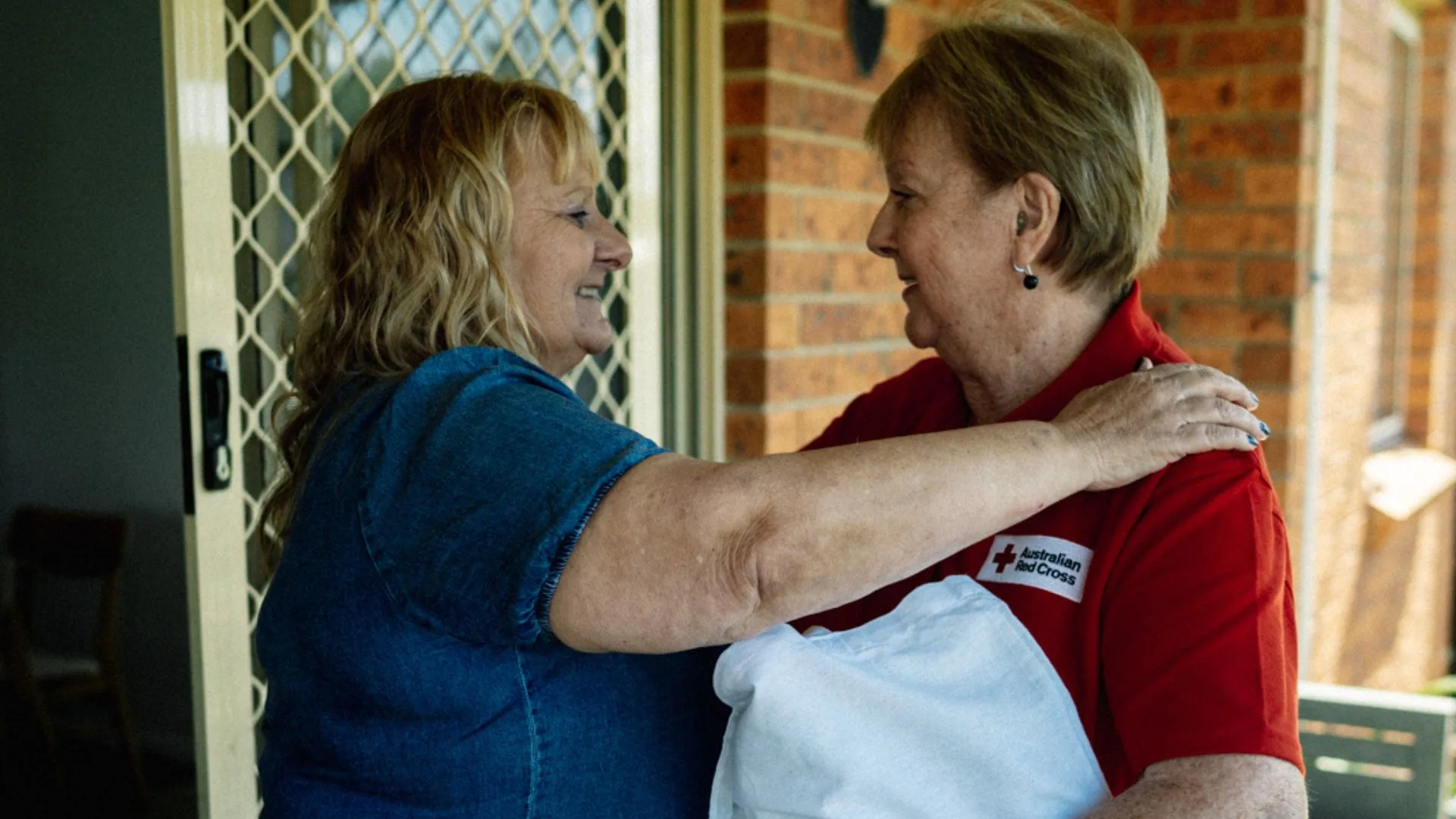 An Australian Red Cross volunteer in a red shirt with the Red Cross logo on her chest smiles and looks at a woman with blonde hair wearing a blue denim-like shirt. The volunteer has her arm around the woman's shoulder, and the woman is looking back at her, also smiling.