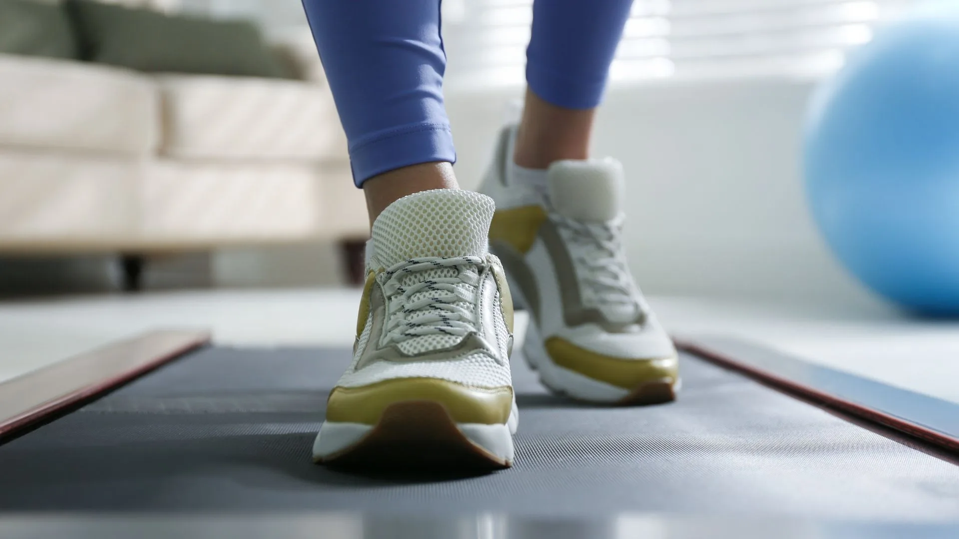 A low-angle shot of a person's legs and feet, wearing white sneakers with gold and tan accents, walking on a dark grey walking pad. The background is a blurred living room with a couch and a large blue exercise ball.