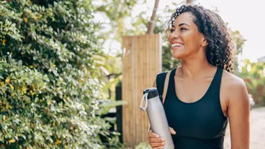 A middle-aged woman with curly hair, wearing a black athletic top, smiles while holding a stainless steel water bottle. She is standing outdoors with lush green foliage and a wooden fence in the background.
