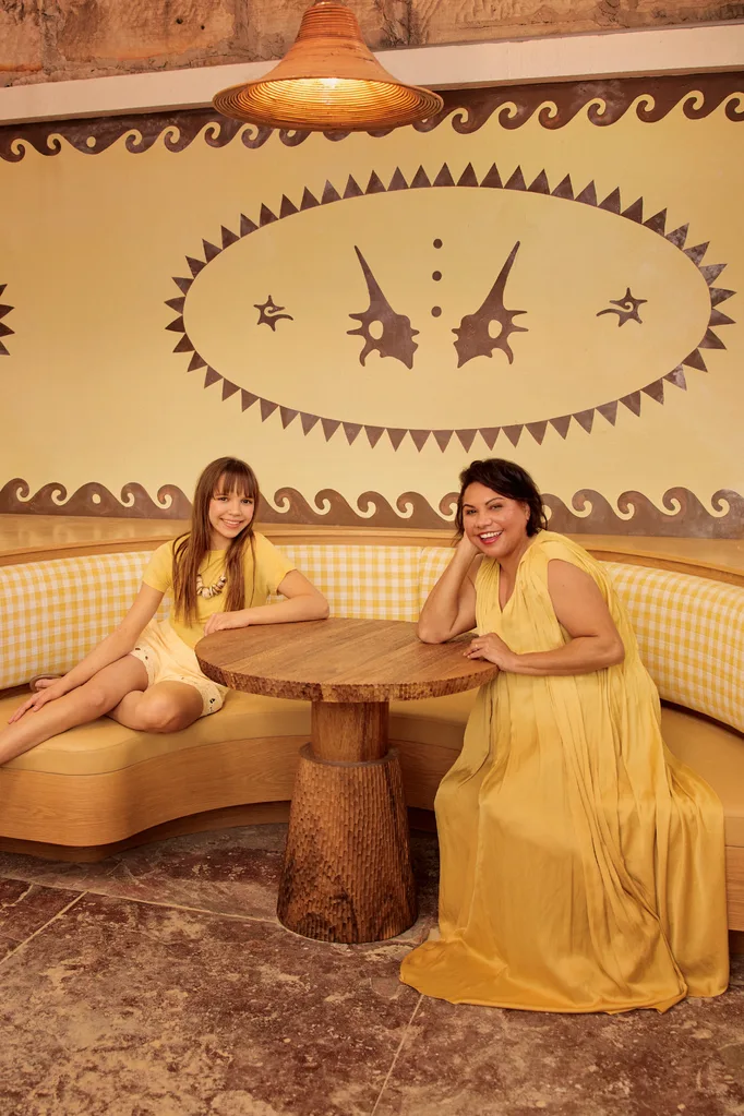 Lily Whiteley and Deborah Mailman wear yellow as they sit by a wooden table.