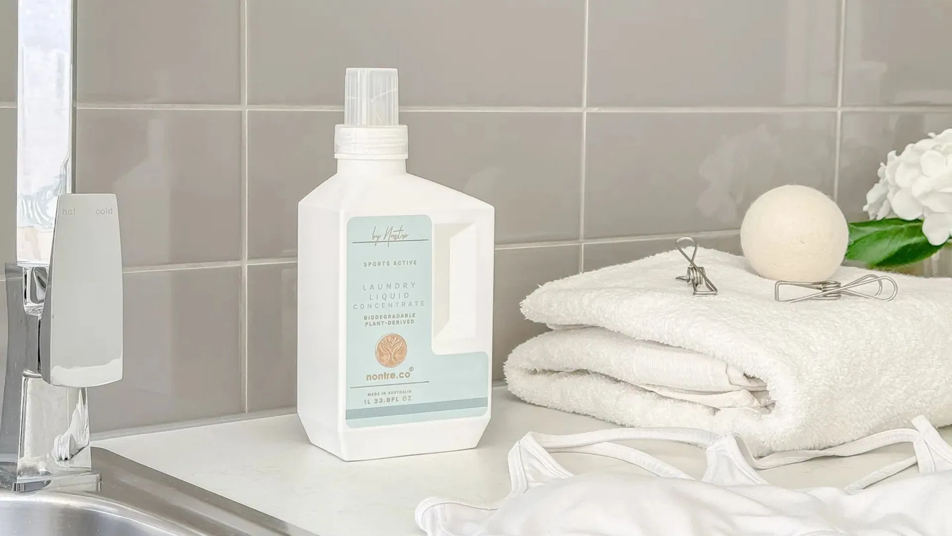 A bottle of Nontre.co Sports Active Laundry Liquid sits on a white countertop next to a stack of clean, white towels, a wool dryer ball, and white activewear. A stainless steel sink faucet is visible to the left.