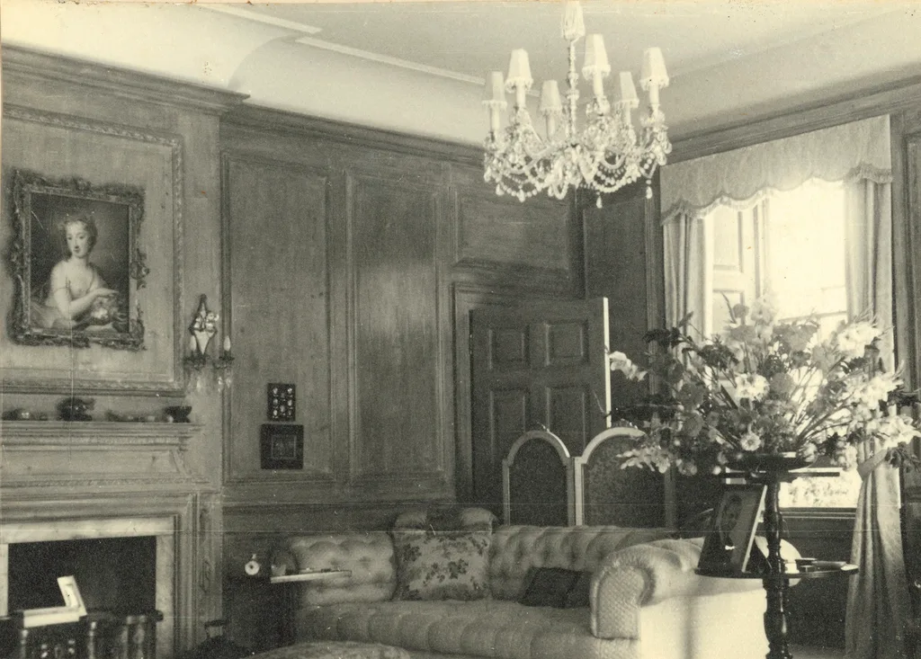 Sepia photo of a sitting room with a couch, vase of flowers, chandelier and a portrait above a fireplace.