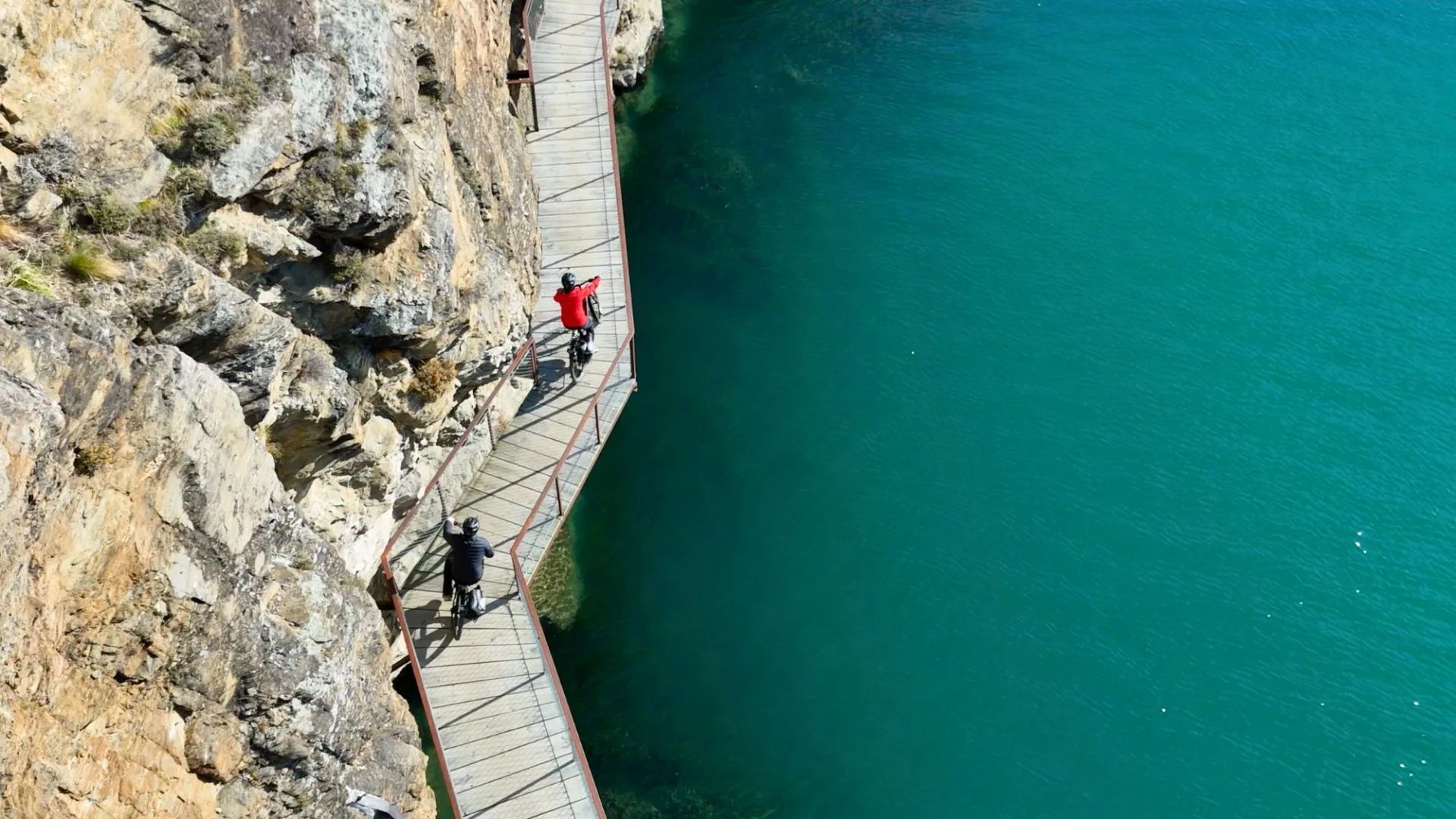 An aerial shot shows two cyclists riding on a boardwalk built directly into a steep rock face. The boardwalk hangs high above a body of vibrant turquoise water, with a metal safety railing on the outer edge.