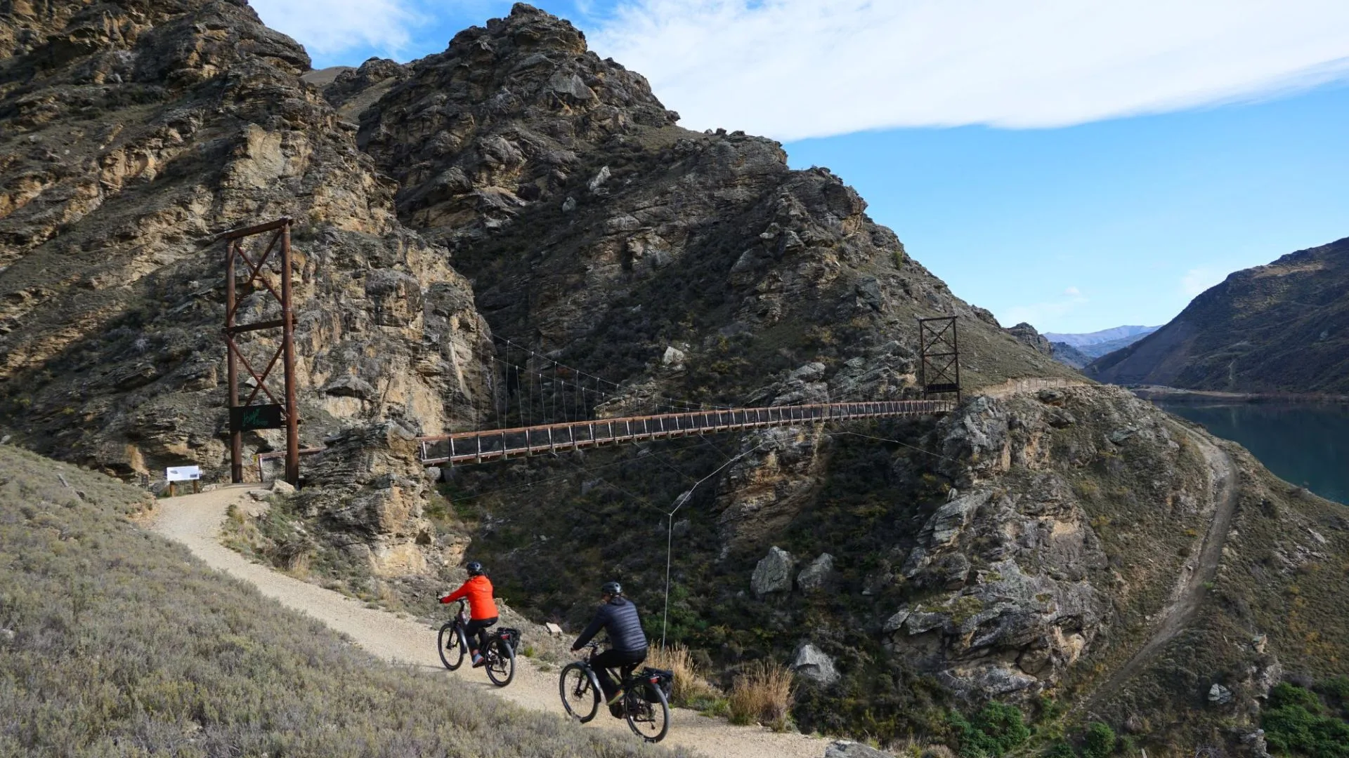 Two cyclists ride on a gravel path winding along a steep, rocky hillside in Central Otago, New Zealand. In the background, a large suspension bridge crosses a deep gorge, connecting the trail across the landscape.