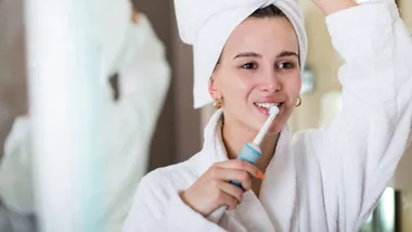 A woman in a white robe and towel uses an electric toothbrush in front of a mirror.