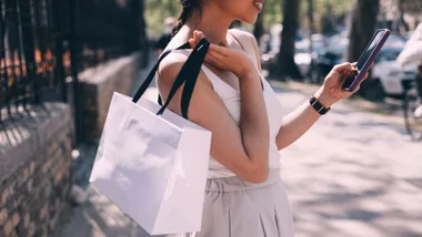 A young woman with a shopping bag and phone smiles while walking outside.