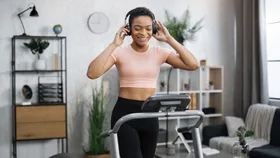 A smiling woman with short hair and headphones walks on a treadmill in a home gym.