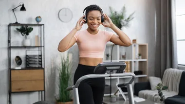 A smiling woman with short hair and headphones walks on a treadmill in a home gym.