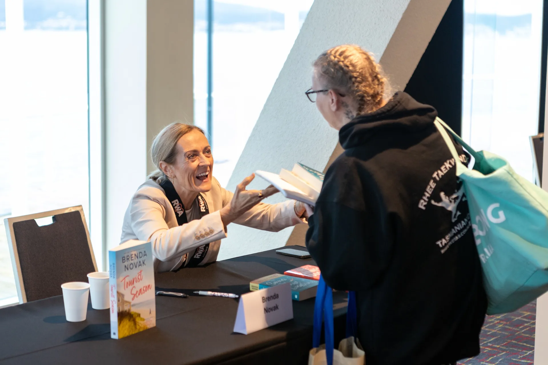 Author Brenda Novak sits at a table, handing a book she has just signed to a woman.