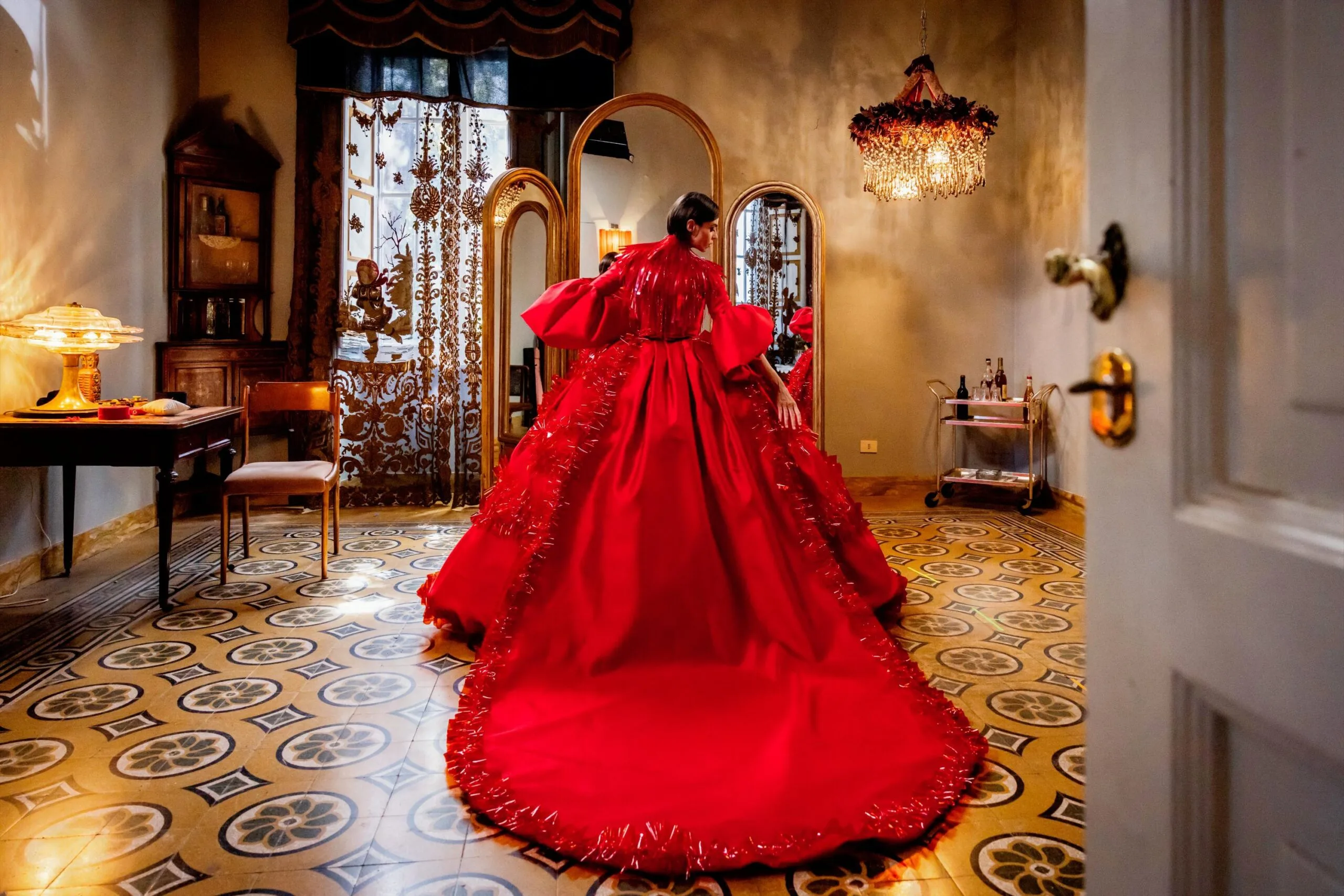 A woman inspects her spectacular red ballgown with a long train in front of the mirror in a stylish tiled apartment.