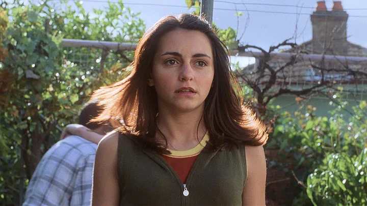 A young woman with black hair looks disconcerted in a backyard garden setting.