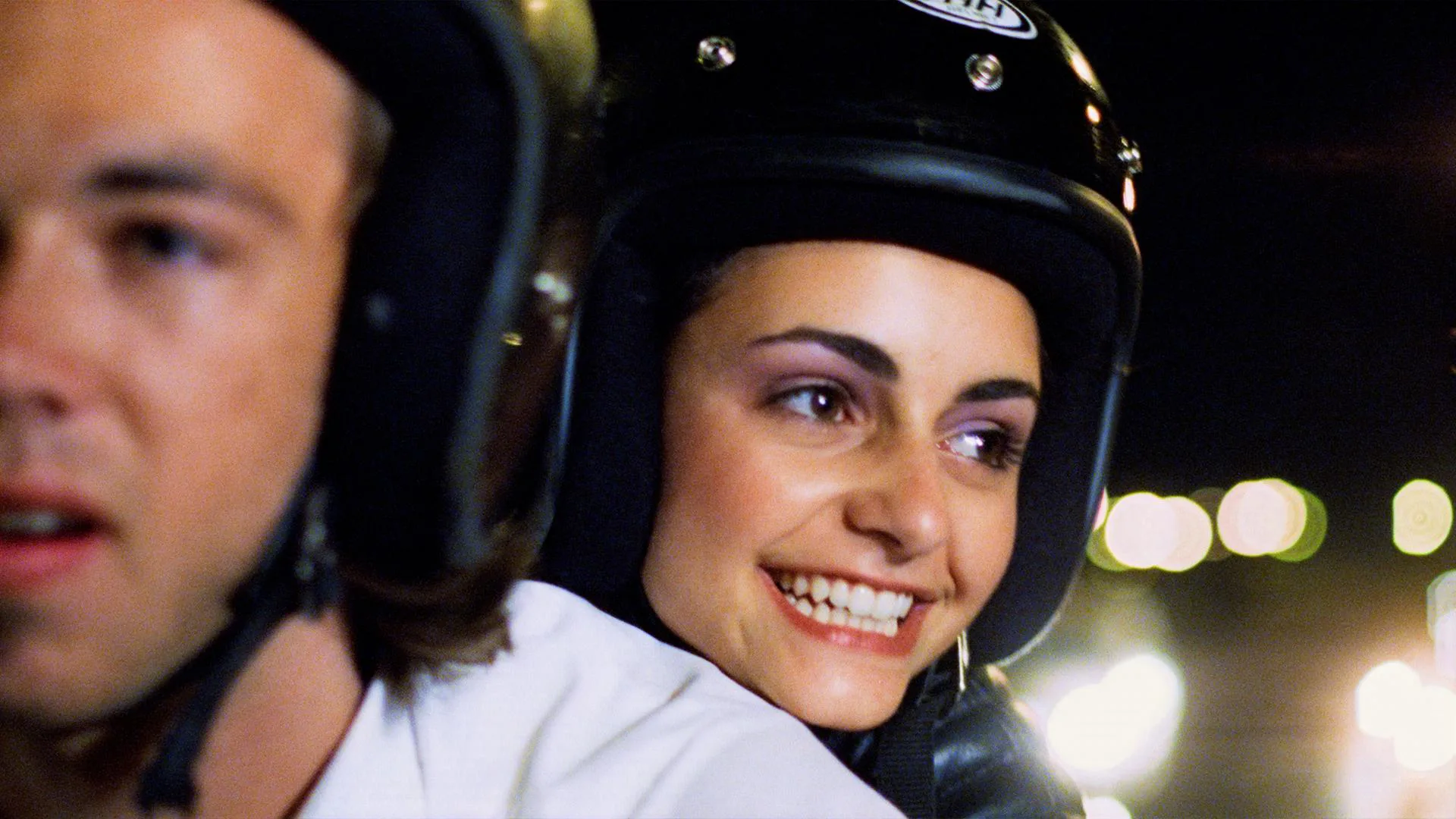 A beaming young woman in a helmet rides on a motorbike behind her male companion