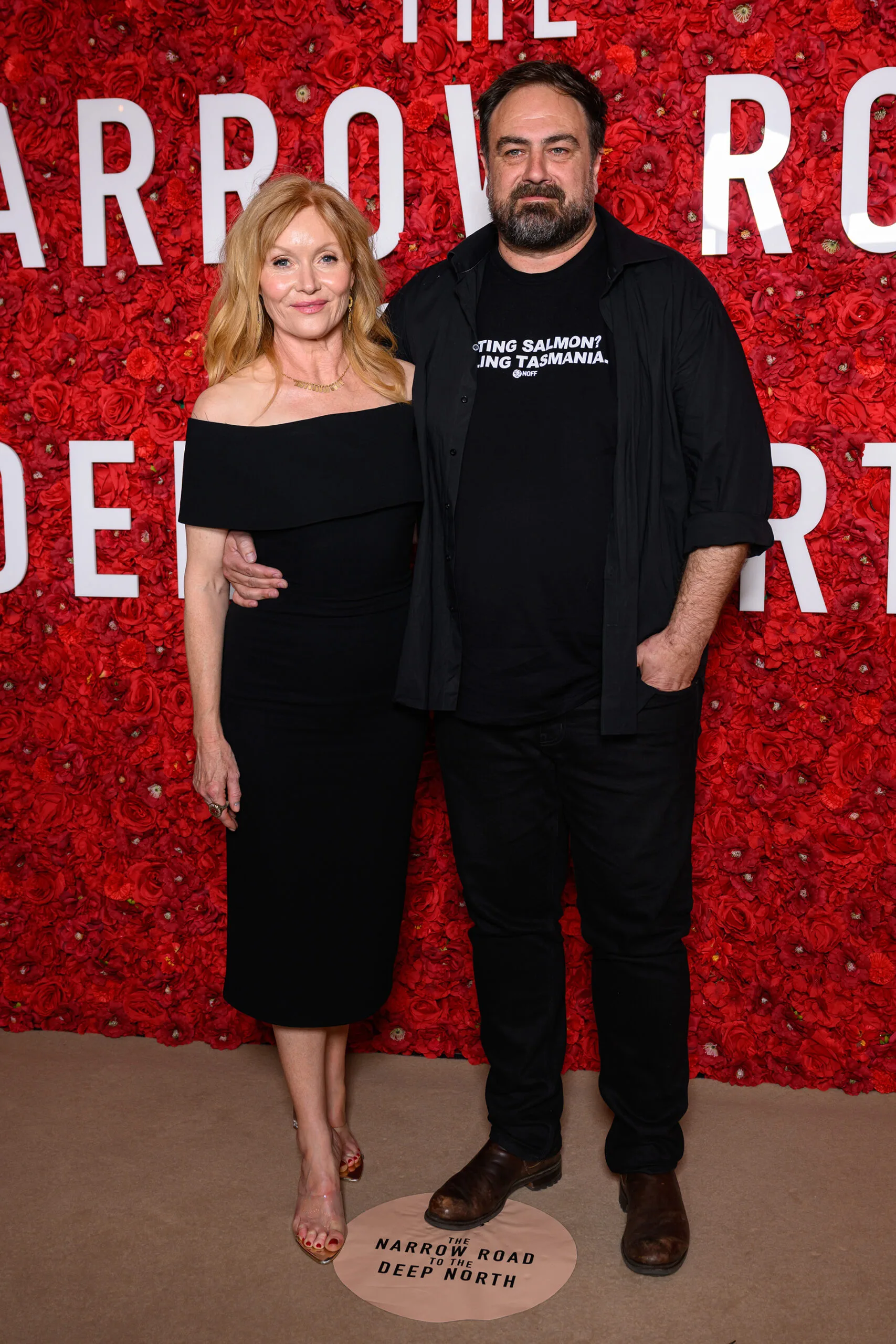 Essie and Justin both wear black. She wears a dress. He wears jeans and an anti-salmon farming t-shirt. The background is a wall of red flowers.