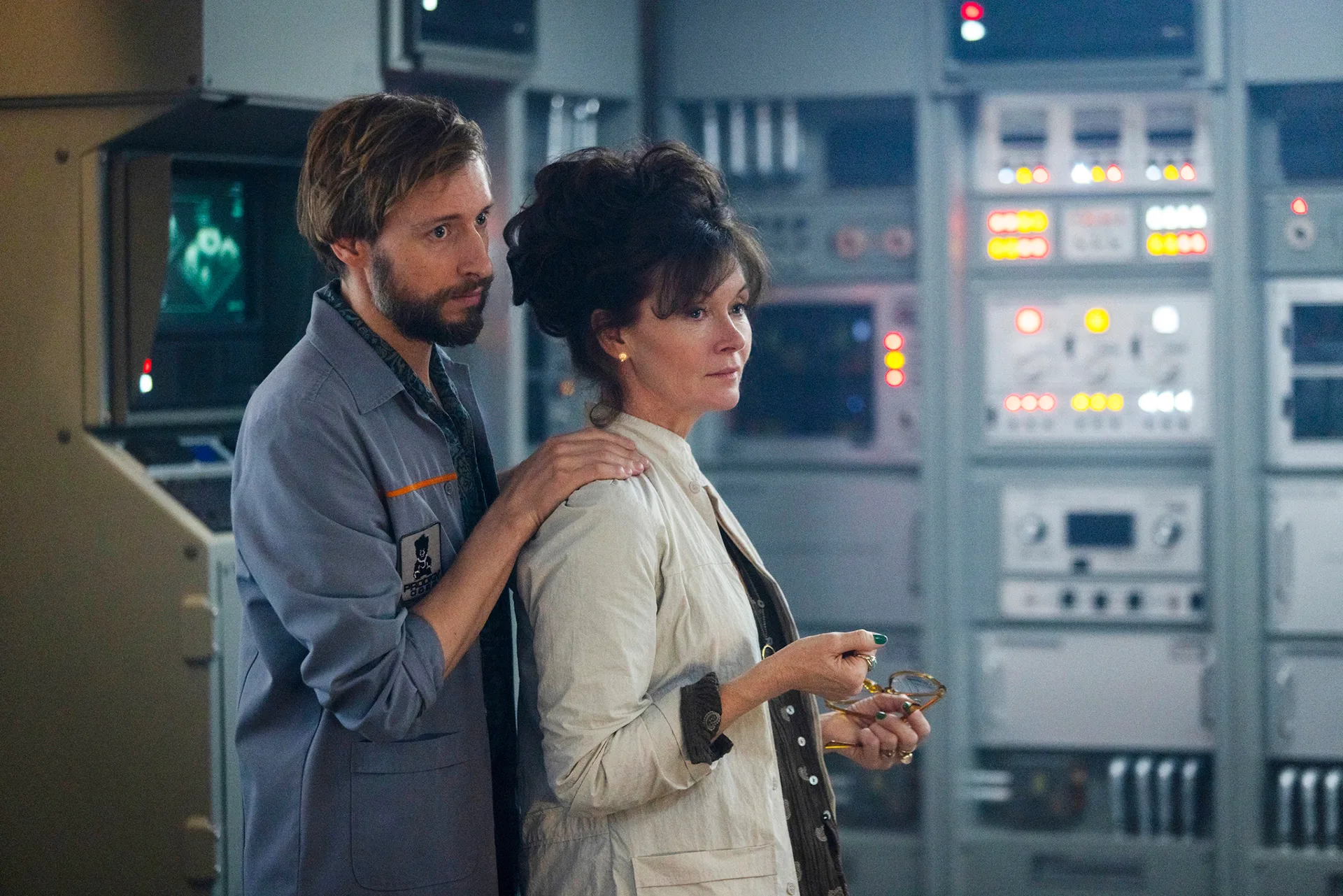 Essie wears a lab coat and her hair is dark and worn in a loose bun. She stands in a scientific lab, with a bearded man behind her, hands on her shoulders.